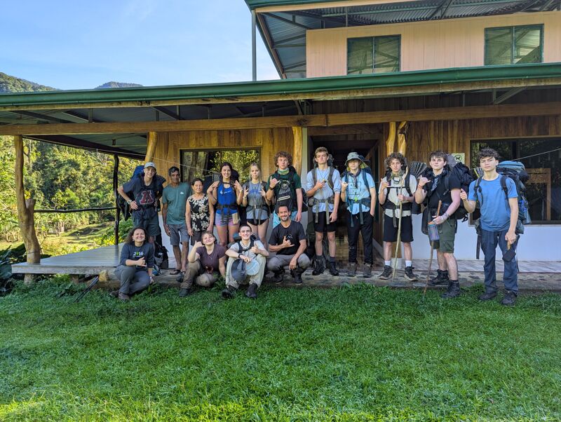 A group of about 18 people are standing outside a building, likely a lodge or hostel, with backpacks. They appear to be on a hiking or backpacking trip. The building has a porch and is surrounded by green grass and trees. The sky is clear and sunny, suggesting good weather for outdoor activities. The group is diverse in age and gender, and they seem to be posing for a photo before or after their adventure.